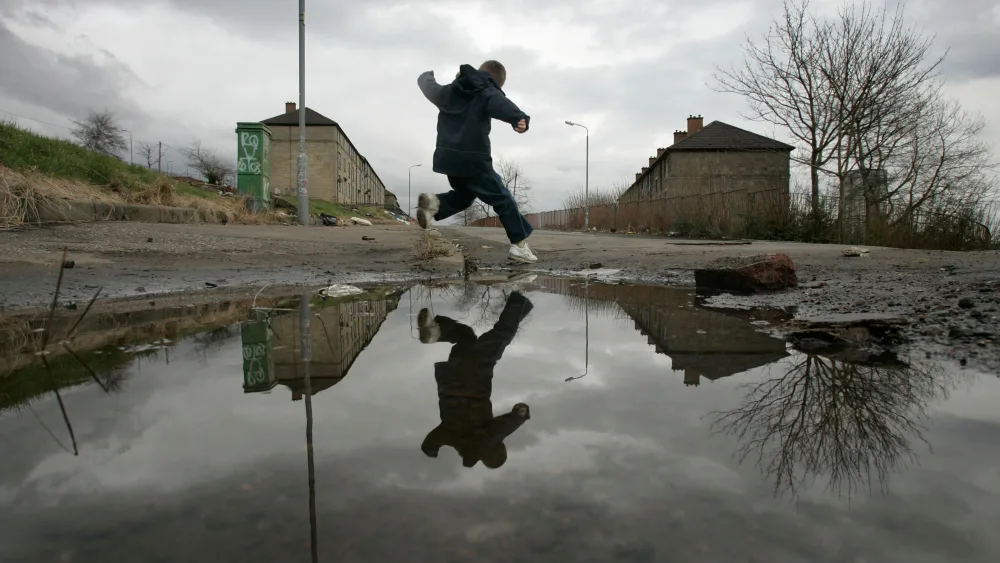 A boy jumping over a large puddle in waste land, with housing in the background