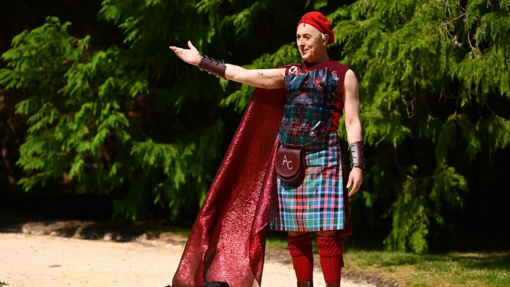 Alan Cumming wearing a tartan outfit, with one arm outstretched