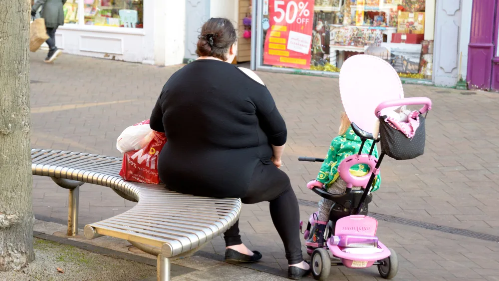 An overweight woman sitting on a bench on a high street, with a small child in a buggy next to her