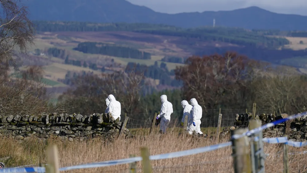 Police in white forensic suits walking in a rural area, with police tape in the foreground
