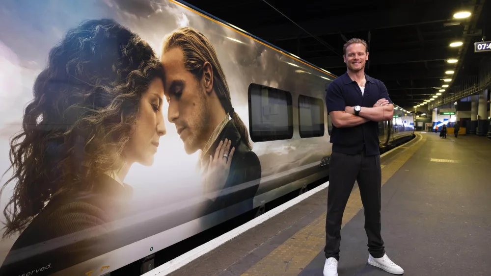 Sam Heughan in front of a train with an Outlander wrap