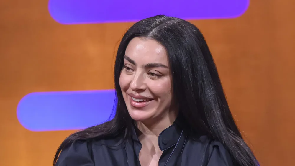 Charli XCX in a leather jacket smiling while sitting on a red couch
