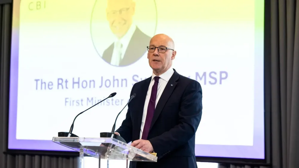 John Swinney speaking from a lectern, with an image of himself projected on the screen behind him