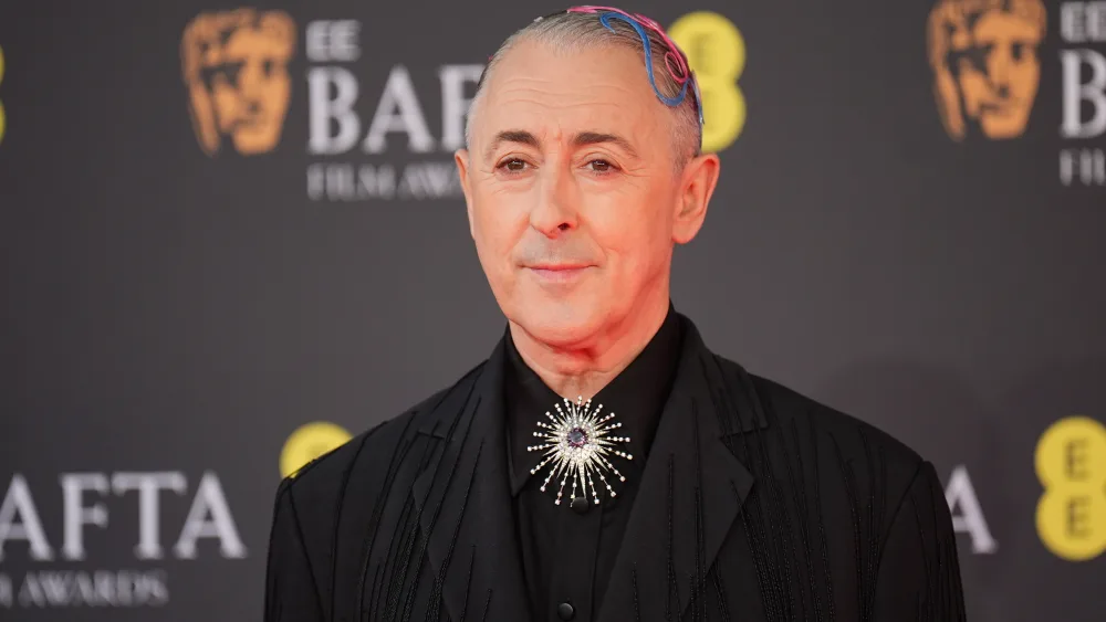 Alan Cumming in a black suit, in front of Bafta Award signage