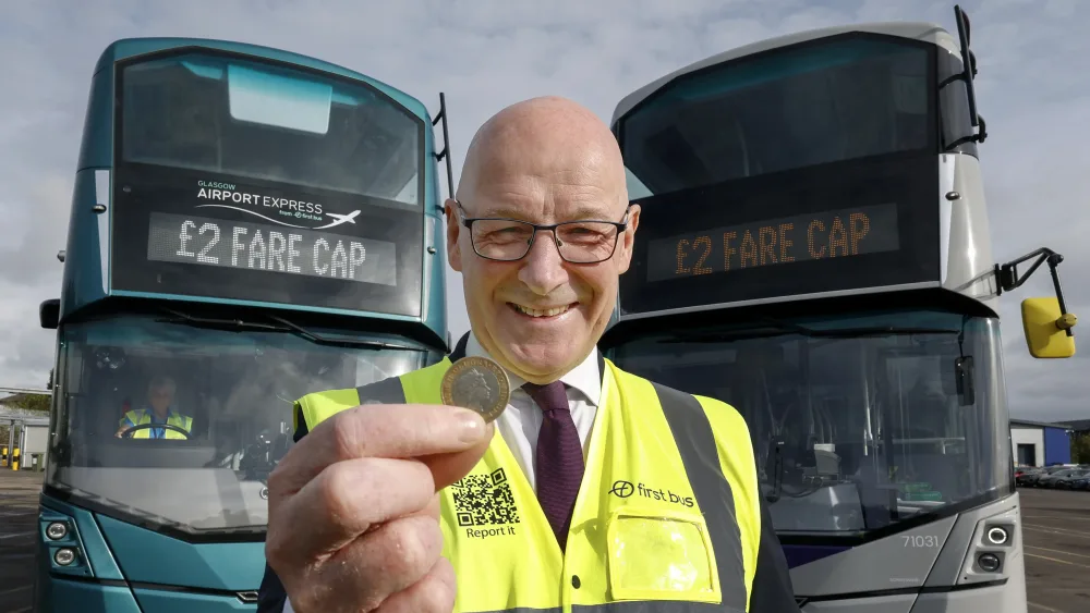 John Swinney smiling while holding up a £2 coin, in front of two buses