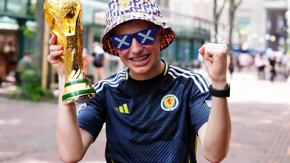 A boy wearing a Scotland top, hat and glasses, holding up a replica World Cup tropy