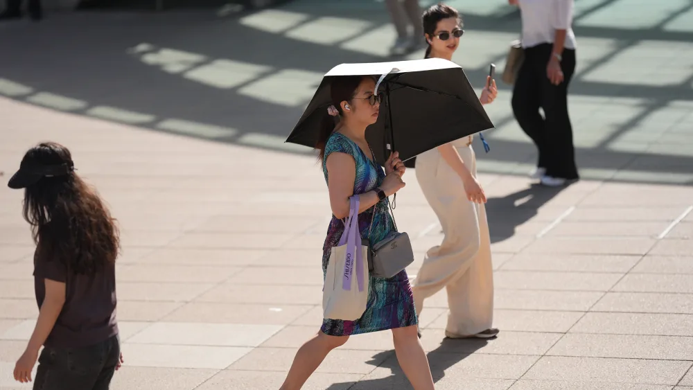 A woman walks across paving in the sunshine sheltering under a black umbrella