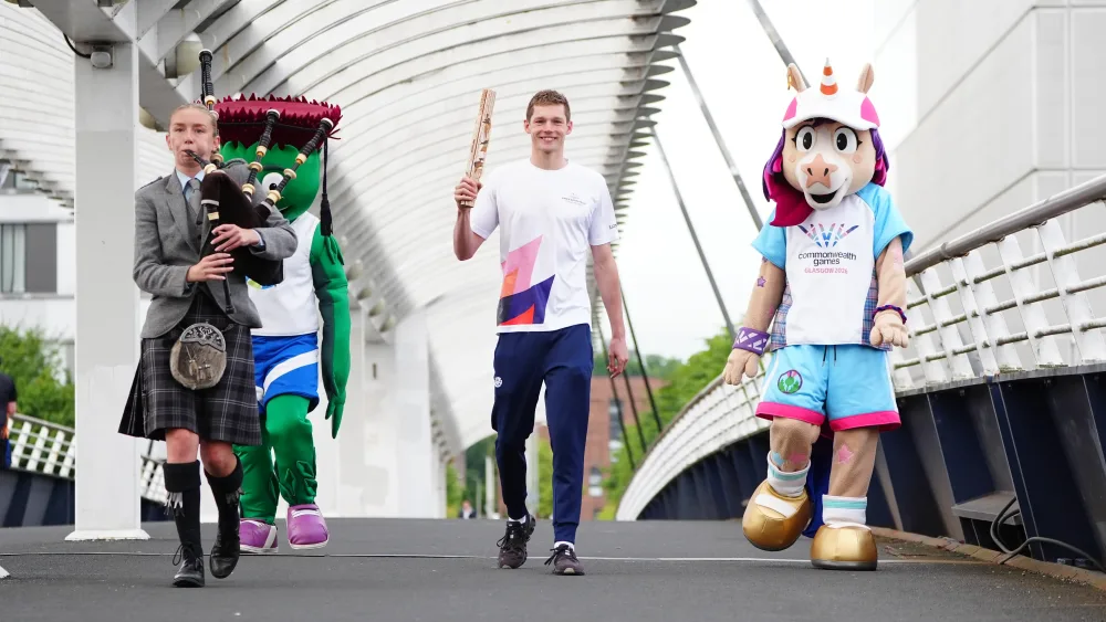 Duncan Scott walks across a bridge holding the King's Baton, flanked by mascots and led by a pipe