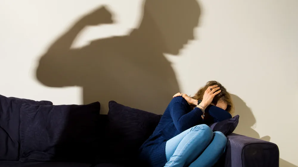 A woman cowering on a sofa, hiding her face, while the shadow of a man looms over her