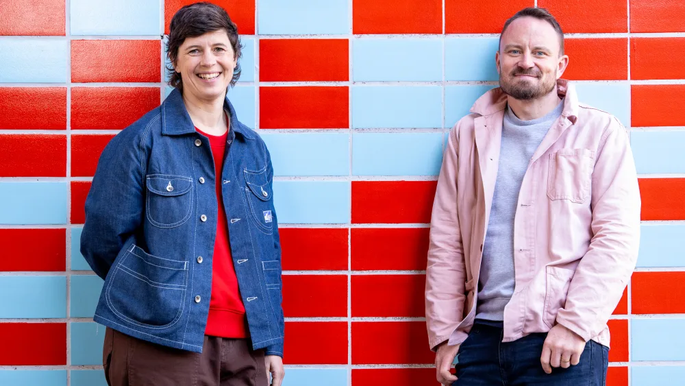 Lucy Osborne and James Grieve smiling, while standing against a tiled, red and blue wall