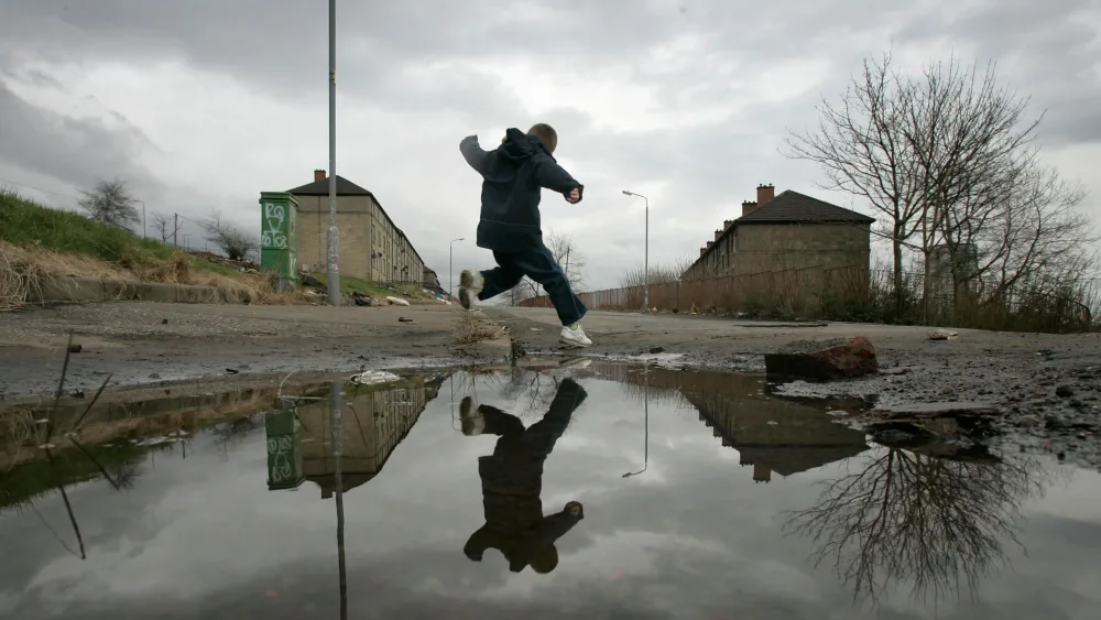 A boy jumping over a large puddle in waste land, with housing in the background