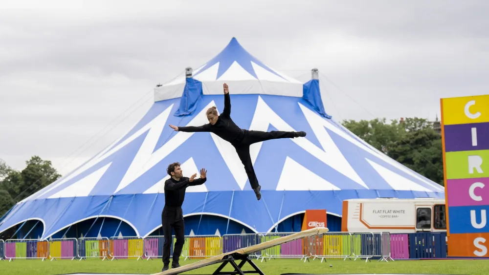 Circus performers, including one jumping in the air, in front of a big blue and white tent