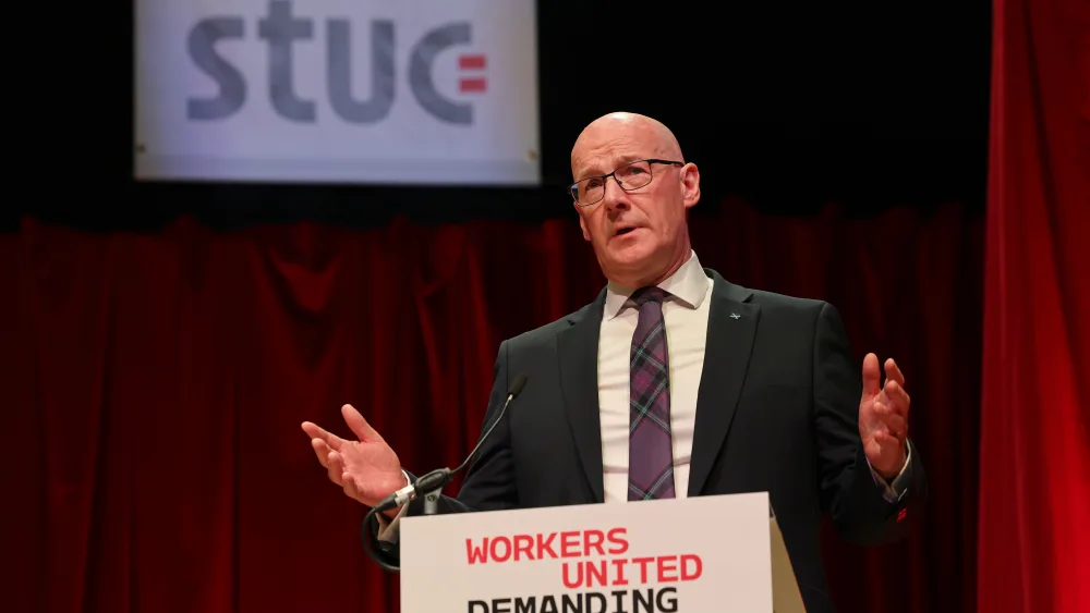 John Swinney speaking on stage, with an STUC sign behind him