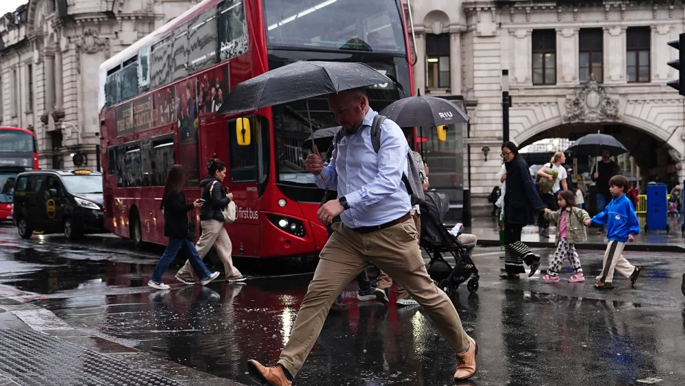 A person crossing a road in the rain, using an umbrella, with a red London bus in the background