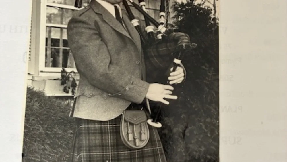 View of a black and white photo of Bruce Williams playing the bagpipes outside a house
